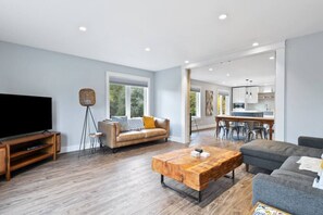 A modern living room with a large flat-screen TV, a wooden coffee table, a sofa, and a dining area with a kitchen in the background. - Skipping Rock Loft: Play, Relax & Recharge (Meaford)