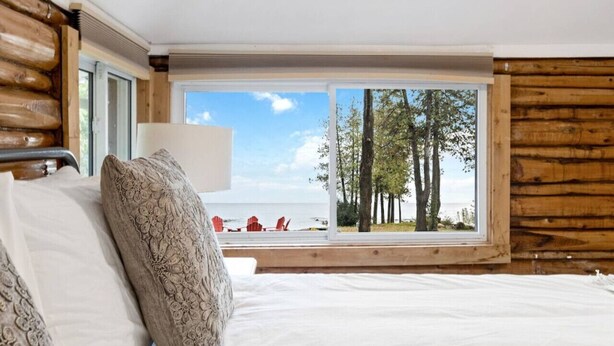 A bedroom with a large window, a bed with patterned pillows, and a view of trees and chairs outside. - Skipping Rock Cottages: Ultimate Lakeside Retreat (Meaford)