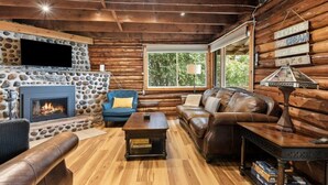 A log cabin interior with a stone fireplace, leather furniture, and a large window offering a view of trees. - Skipping Rock Cottages: Ultimate Lakeside Retreat (Meaford)