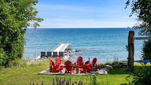 A coastal view with a pier, red Adirondack chairs, and a grassy area with purple flowers. - Skipping Rock Cottages: Ultimate Lakeside Retreat (Meaford)