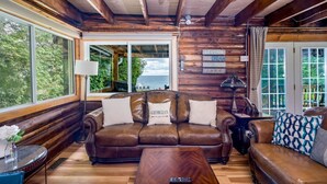 A cozy cabin interior with a leather sofa, wooden walls, and a large window overlooking a scenic view. - Skipping Rock Cottages: Ultimate Lakeside Retreat (Meaford)