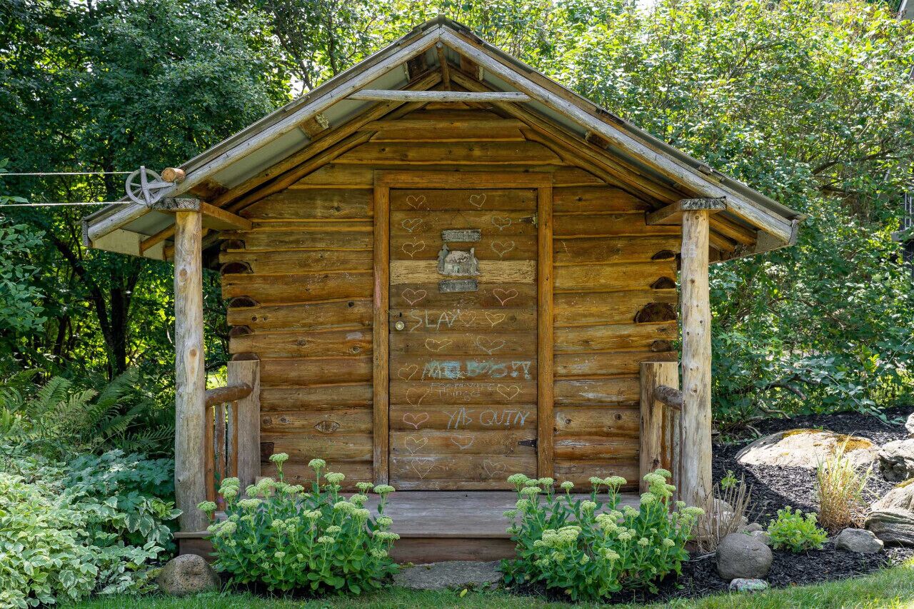 A rustic wooden cabin with a front porch, surrounded by lush greenery.