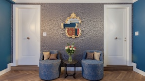 A room with two blue upholstered chairs, a small table with a vase of flowers, and a decorative mirror on the wall. - 286 Piazza San Marco (Venice)