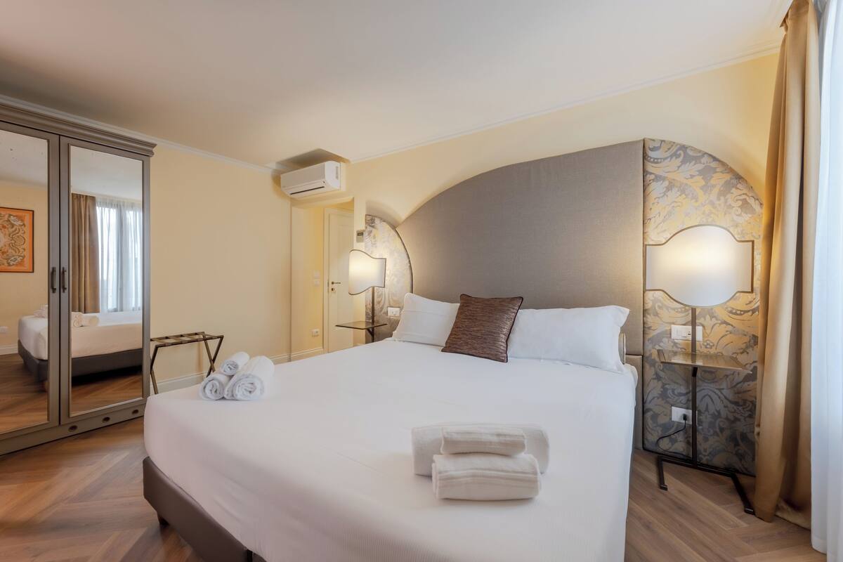 A hotel room with a large bed, a bedside table with a lamp, a mirror, and a wardrobe.