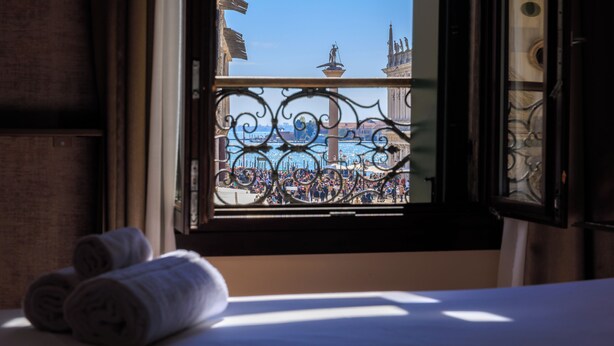 A hotel room with a bed, a window with decorative iron railings, and a view of a cityscape with buildings and a statue. - 286 Piazza San Marco (Venice)