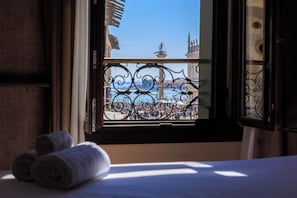 A hotel room with a bed, a window with decorative iron railings, and a view of a cityscape with buildings and a statue. - 286 Piazza San Marco (Venice)