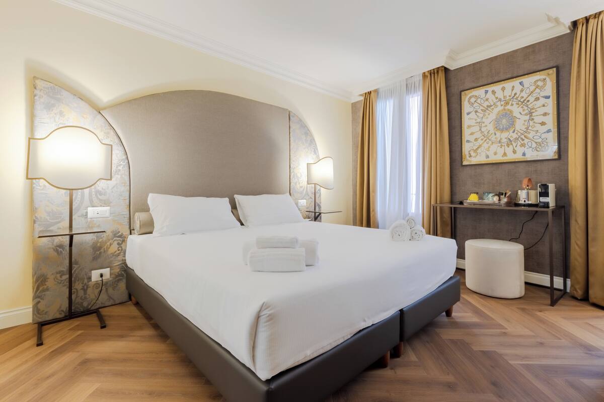 A large bed with white linens, two bedside tables with lamps, a small ottoman, and a decorative wall piece.