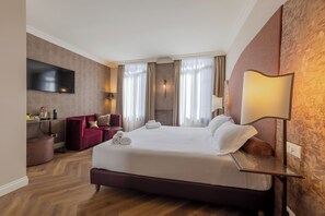 A hotel room with a large bed, a desk with a flat-screen TV, a red armchair, and a wall-mounted lamp.