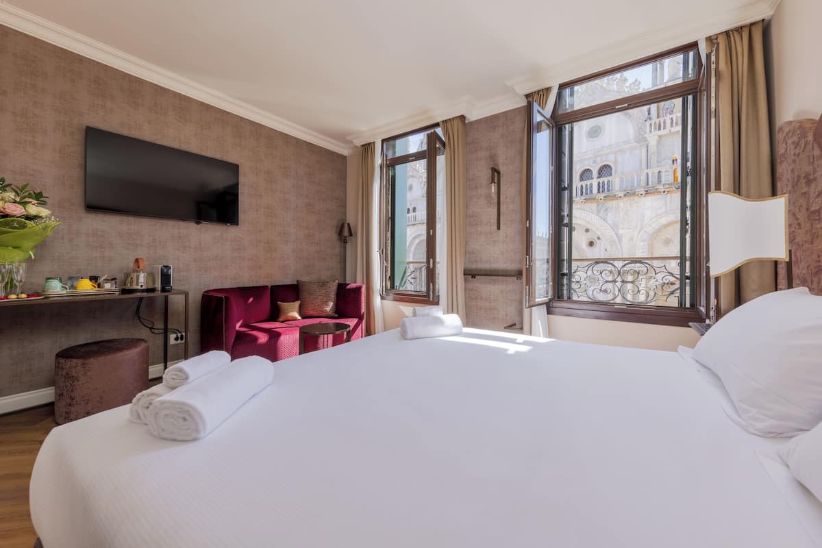 A hotel room with a large bed, a TV mounted on the wall, a desk with a chair, and a view of a historic building through the window.