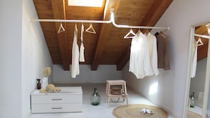 Quarto com teto inclinado, um cabideiro suspenso, um pequeno banquinho de madeira, um tapete redondo de fibra natural e um espelho refletindo o espaço.