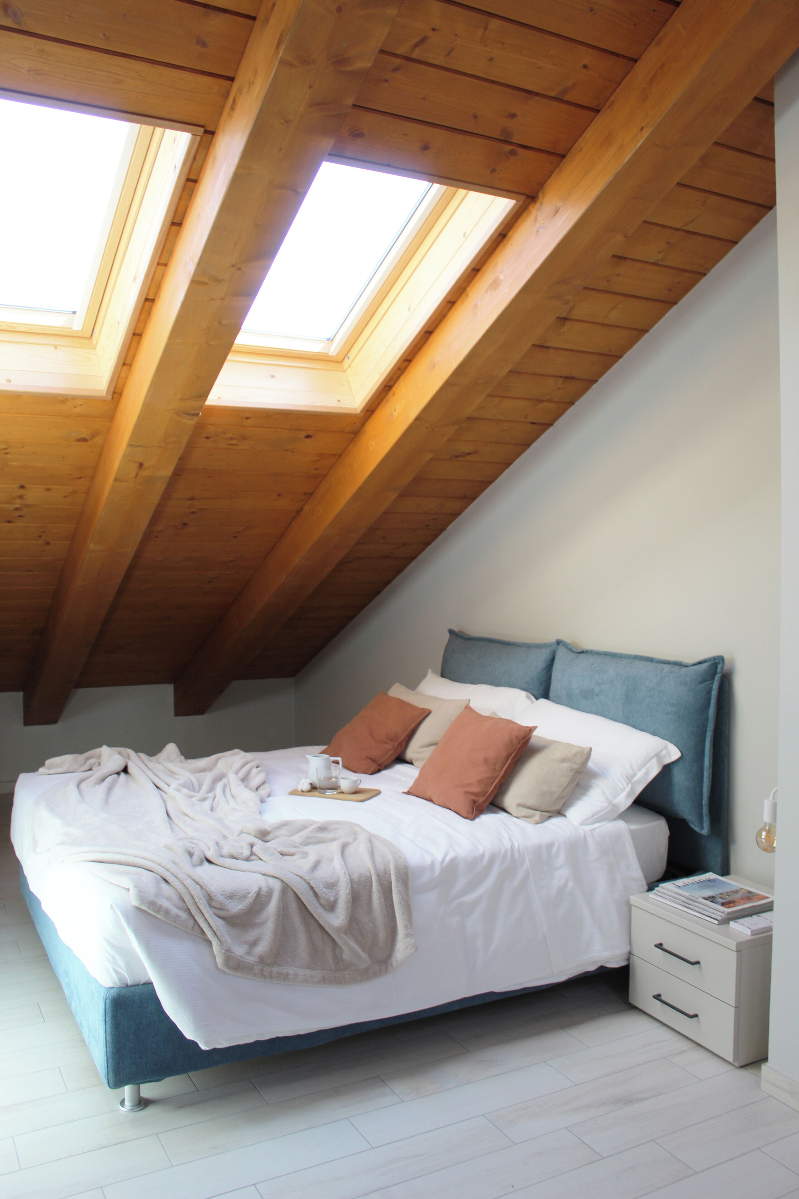 A bedroom with a sloped ceiling, skylights, a bed with pillows, a bedside table, and a nightstand.