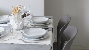 Uma mesa de jantar posta com pratos, copos e talheres, com um vaso de flores secas e uma vela em uma prateleira acima.