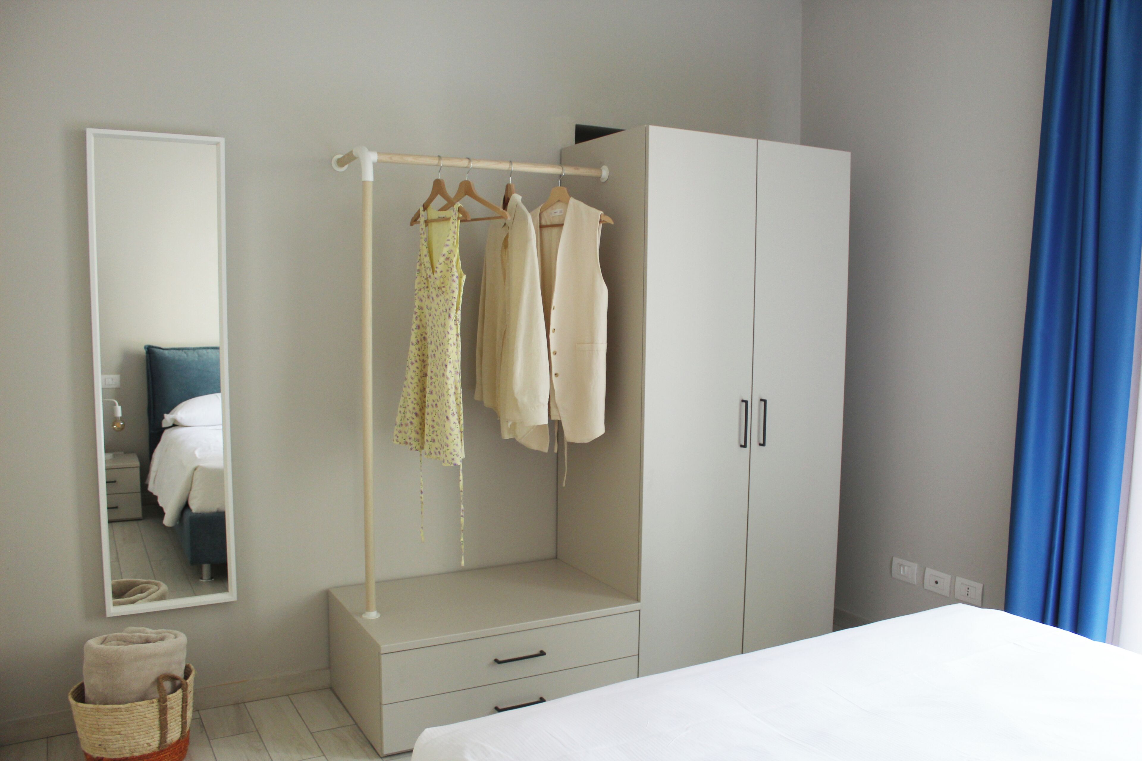 A bedroom with a bed, a wardrobe, a hanging clothes rack, a mirror, and a small basket with towels.