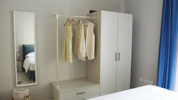 A bedroom with a bed, a wardrobe, a hanging clothes rack, a mirror, and a small basket with towels.