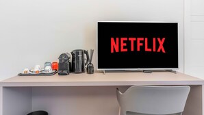 A modern workspace with a flat-screen TV displaying "NETFLIX" on the screen, a coffee maker, a kettle, and a mug on the desk.
