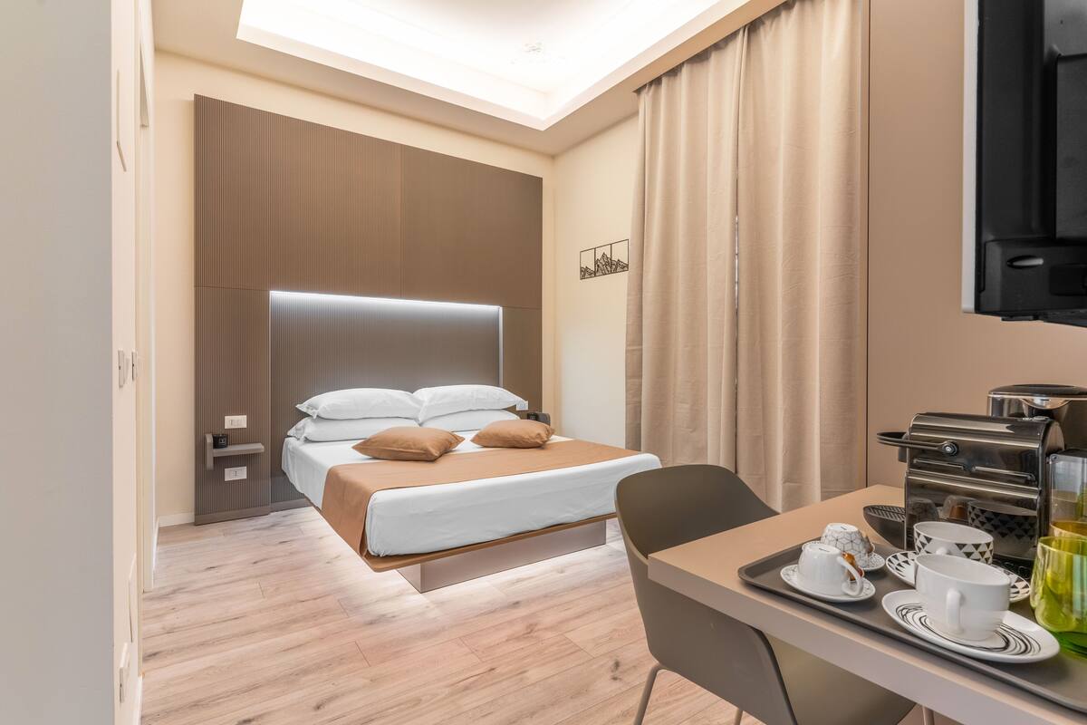 A modern hotel room with a bed, bedside tables, a desk with a coffee maker, and a TV.