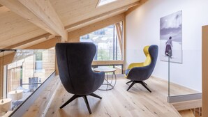 Un interno moderno con soffitto a falde, travi in legno e ampie finestre che offrono vista sull'esterno. Nell'ambiente, due poltrone e un tavolino.