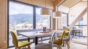 Zona pranzo con tavolo e sedie in legno, ampie finestre che offrono vista su case e montagne, e lampade a sospensione.