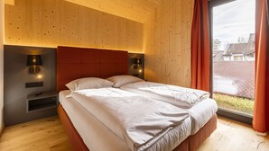 A modern bedroom with a wooden ceiling, a large bed with white bedding, two bedside lamps, and a view of the outside through a large window. - Chalet # 5a with sauna & pool (St. Georgen am Kreischberg)
