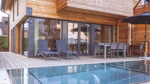 A modern outdoor pool area with lounge chairs and a table, surrounded by wooden deck and glass doors. - Chalet # 5a with sauna & pool (St. Georgen am Kreischberg)