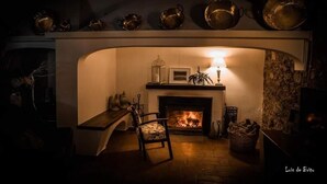 Une pièce chaleureuse avec une cheminée, un mur en pierre, une chaise en bois et une table avec une lampe.