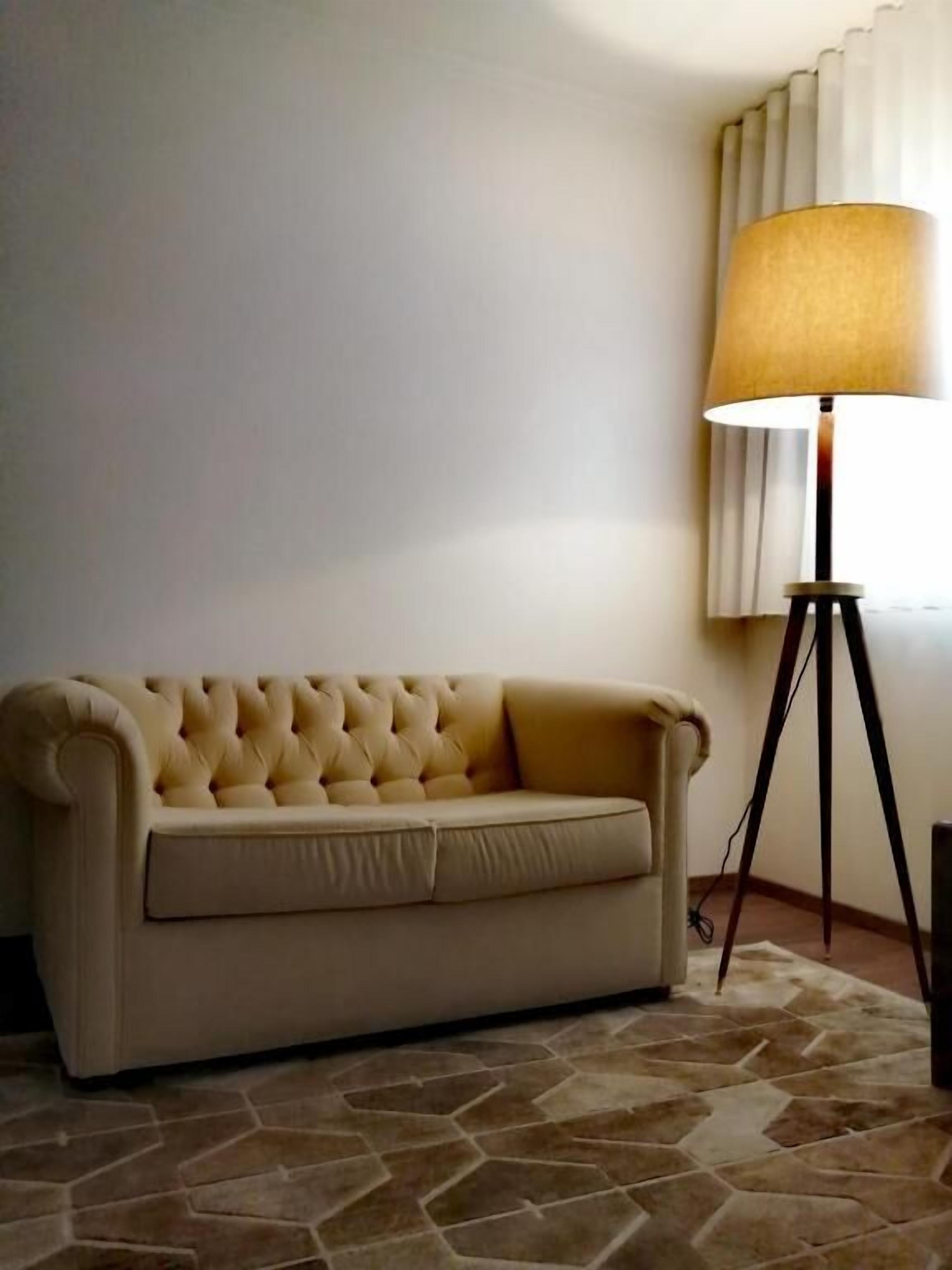 A beige tufted sofa against a plain wall, a floor lamp with a beige lampshade, and a stone-patterned floor.