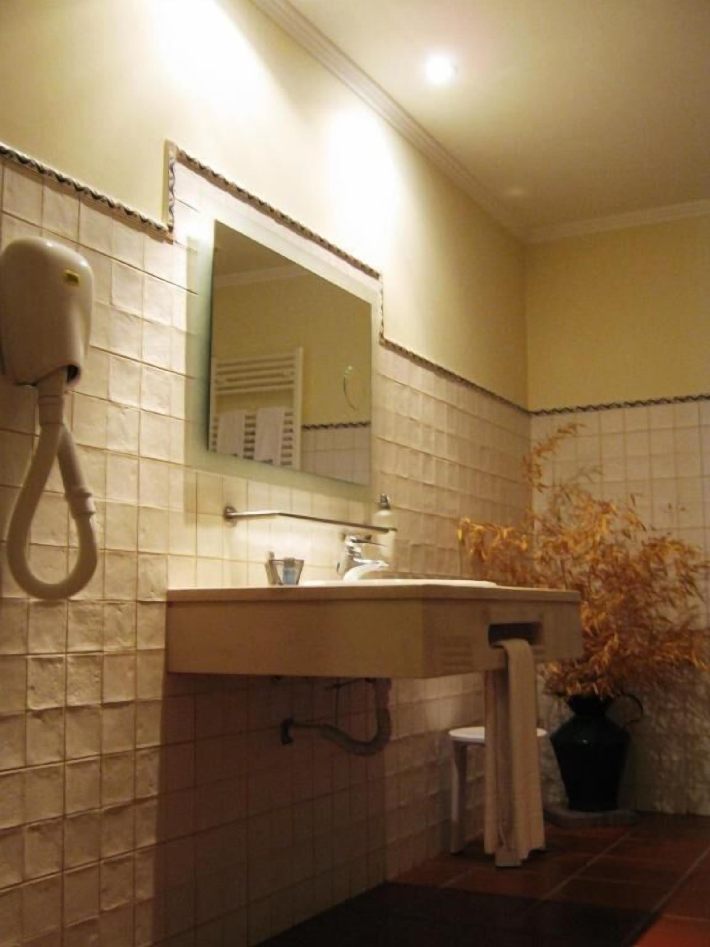 Une salle de bain avec un mur recouvert de carreaux blancs, un lavabo avec un miroir au-dessus, un sèche-cheveux et une plante en pot.