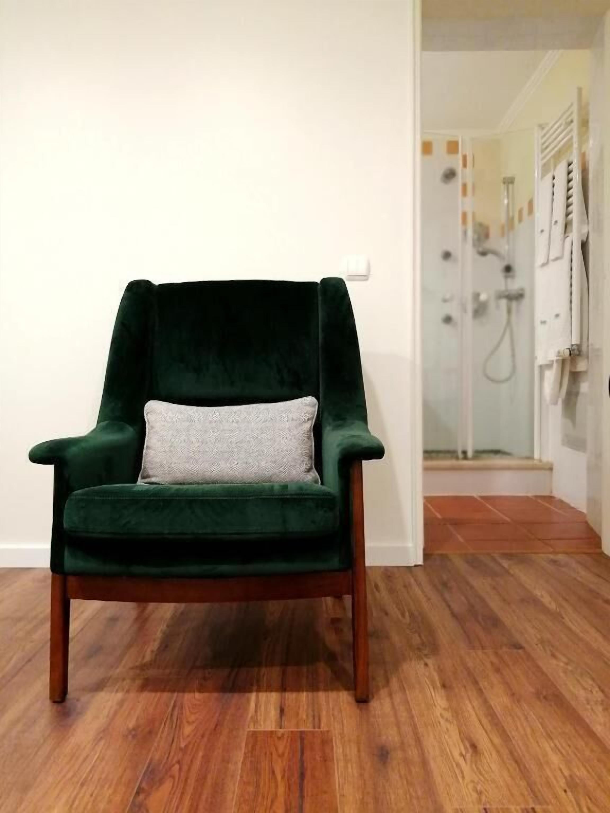 A dark green velvet armchair with a light grey cushion in a room with wooden flooring and a glass shower door in the background.