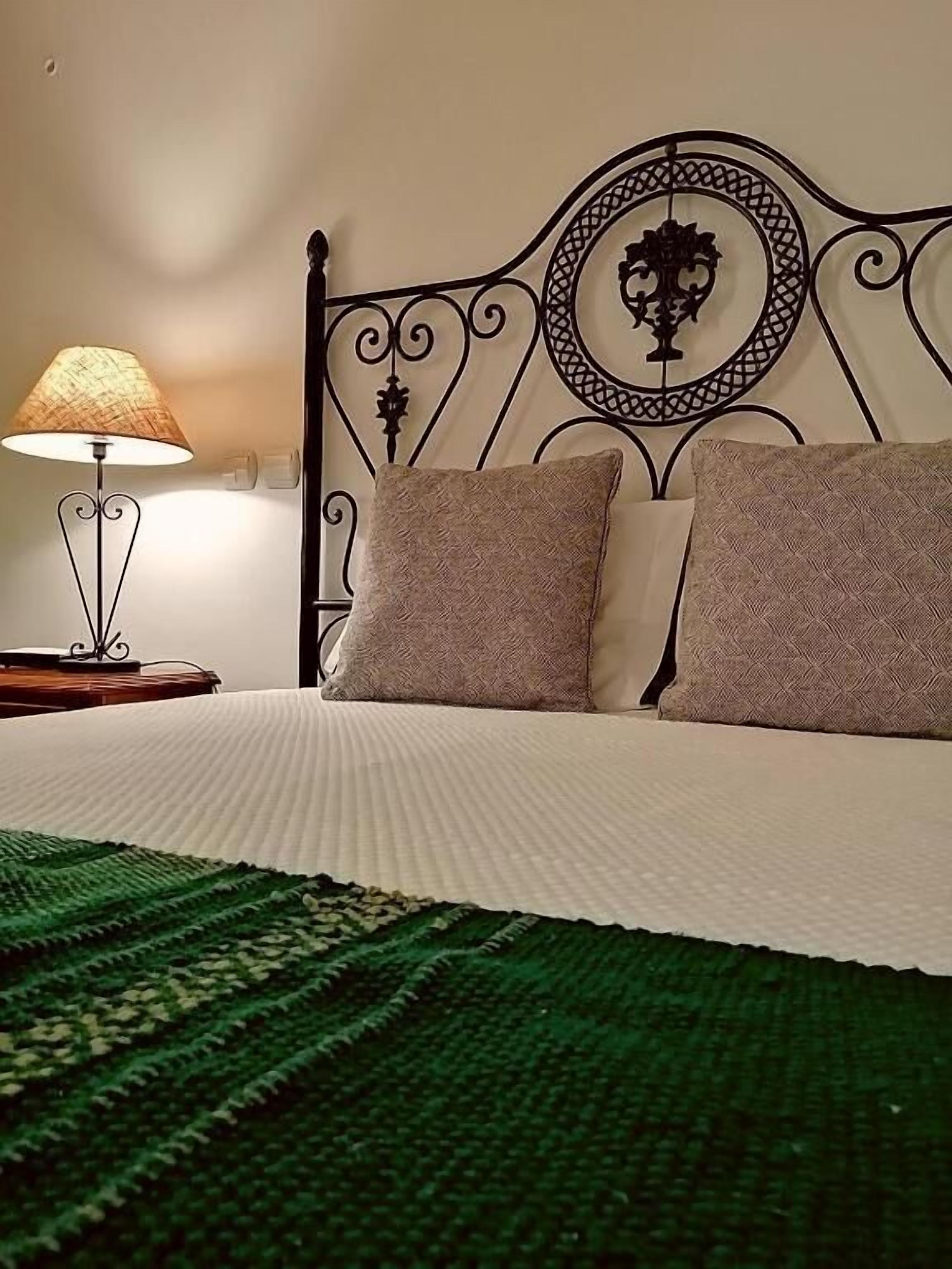 A neatly made bed with a patterned green blanket, a decorative metal headboard, and a bedside lamp.