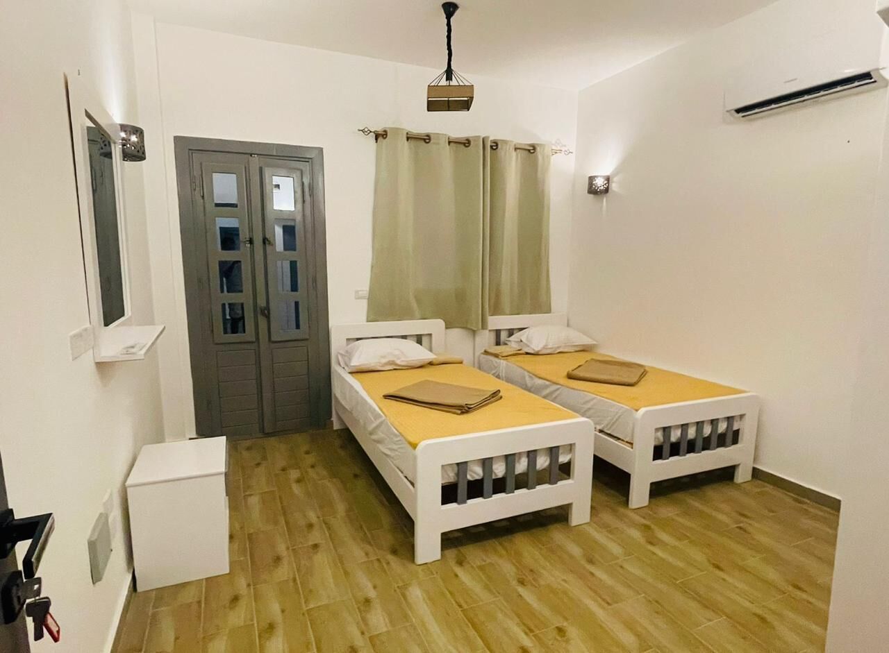 A room with two single beds, a wooden floor, a door with glass panels, a small white cabinet, and a ceiling light fixture.