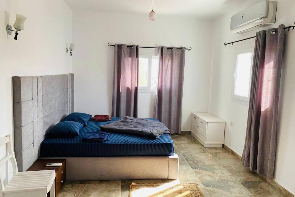 A bedroom with a bed, a bedside table, a chair, a window with curtains, and an air conditioning unit.
