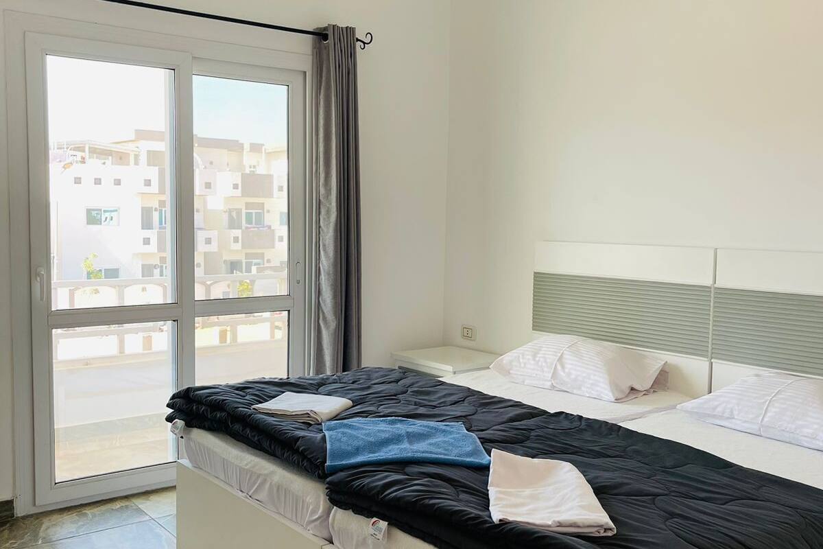 A bedroom with a bed, a window with curtains, a ceiling air conditioner, and a view of buildings outside.