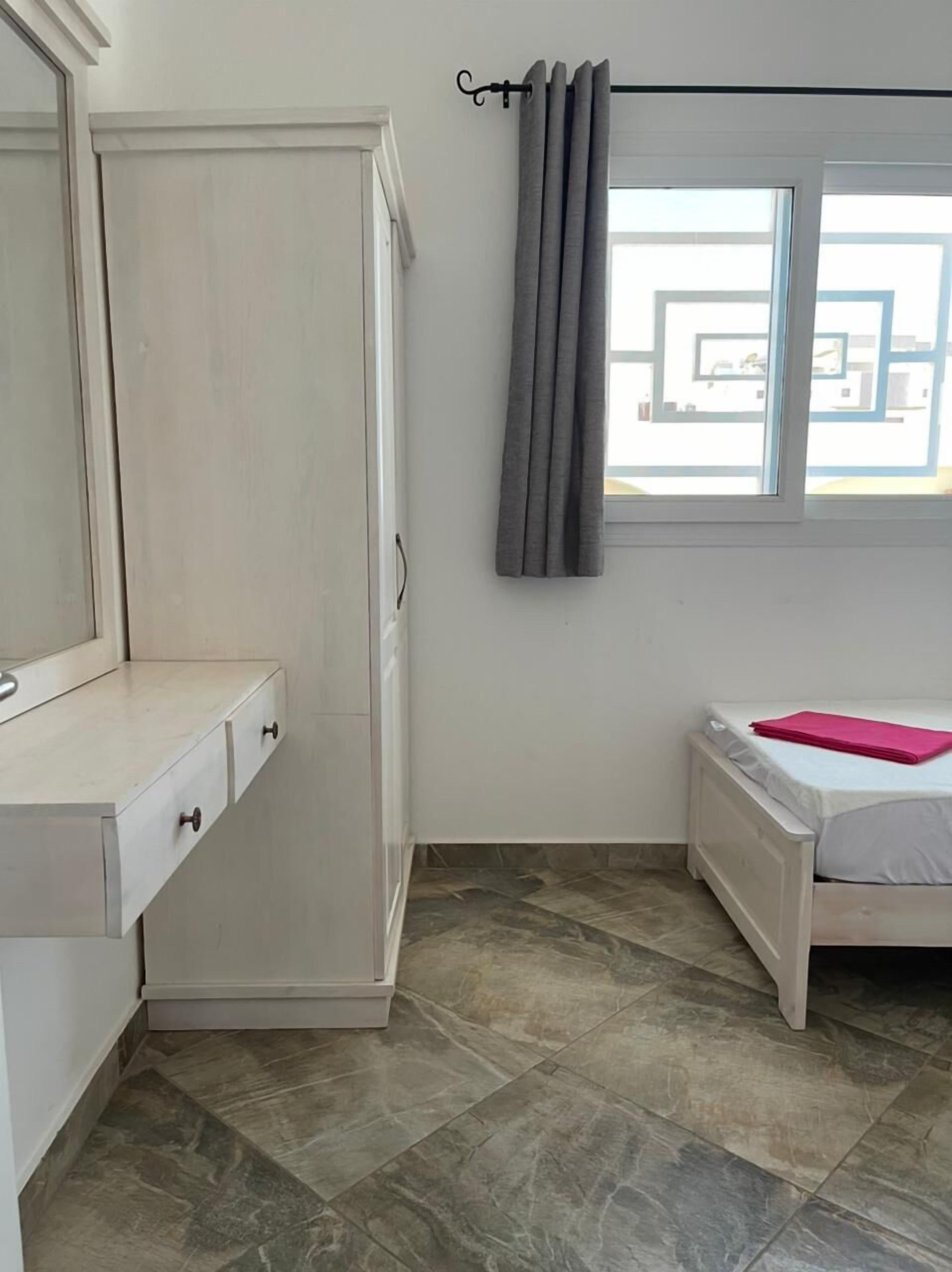 Quarto com guarda-roupa branco, mesa de cabeceira, cama com manta rosa e janela com cortinas.
