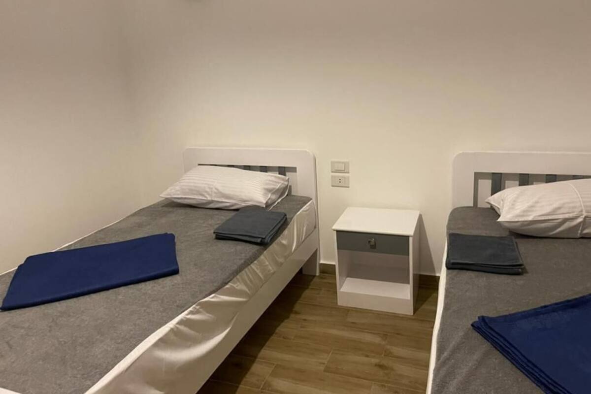 A room with two single beds, each with a pillow and a folded blanket. There is a small white nightstand between the beds.