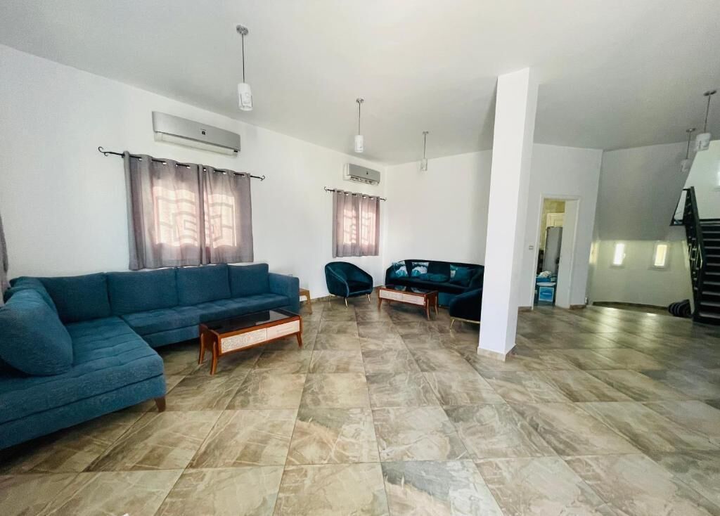 A spacious living room with a sectional sofa, a coffee table, and a TV. There are two windows with curtains, an air conditioning unit, and a staircase in the background.