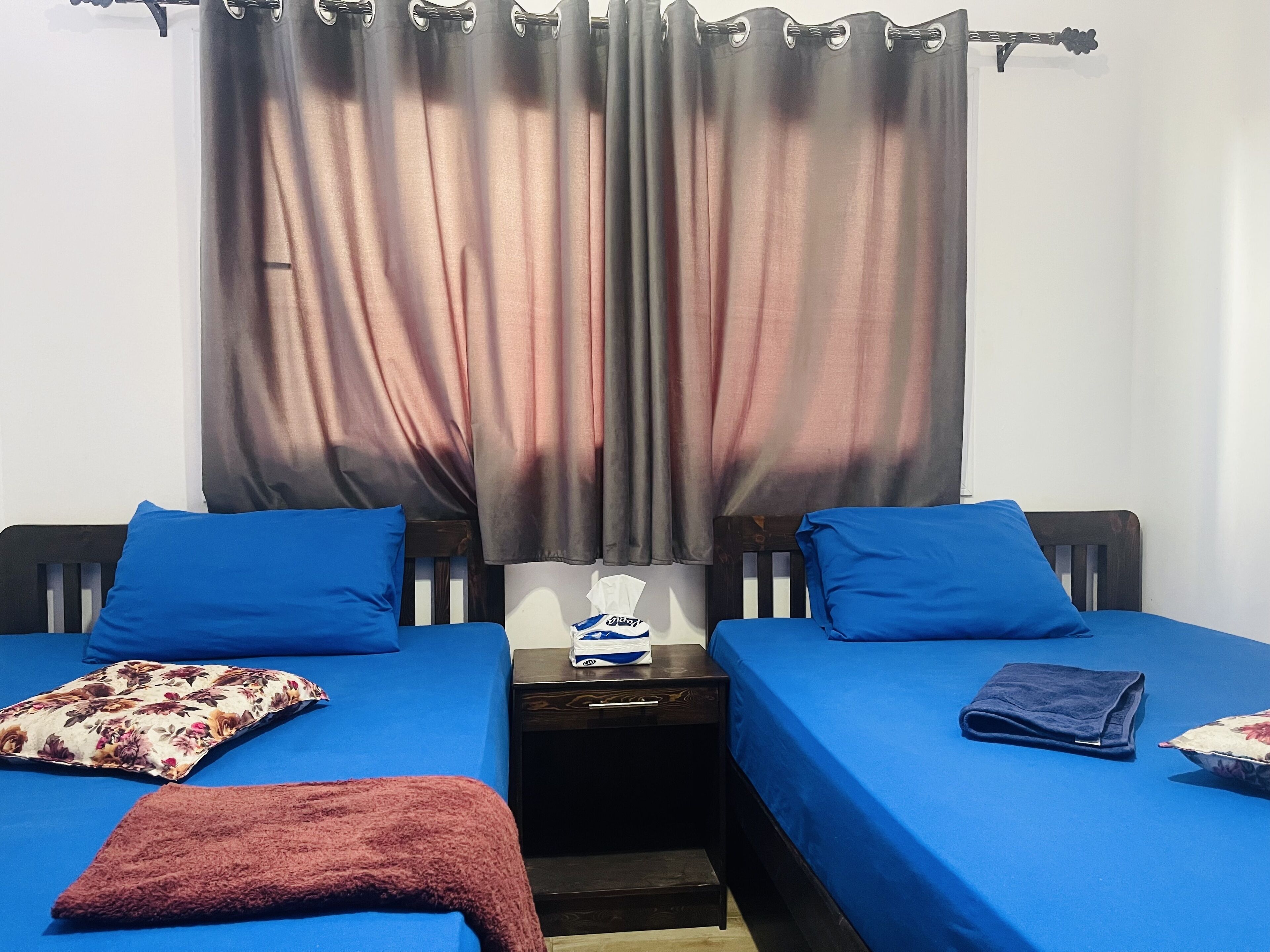 A room with two beds, each with a blue bedspread and matching pillows. There is a small table with a folded towel and a pillow between the beds.