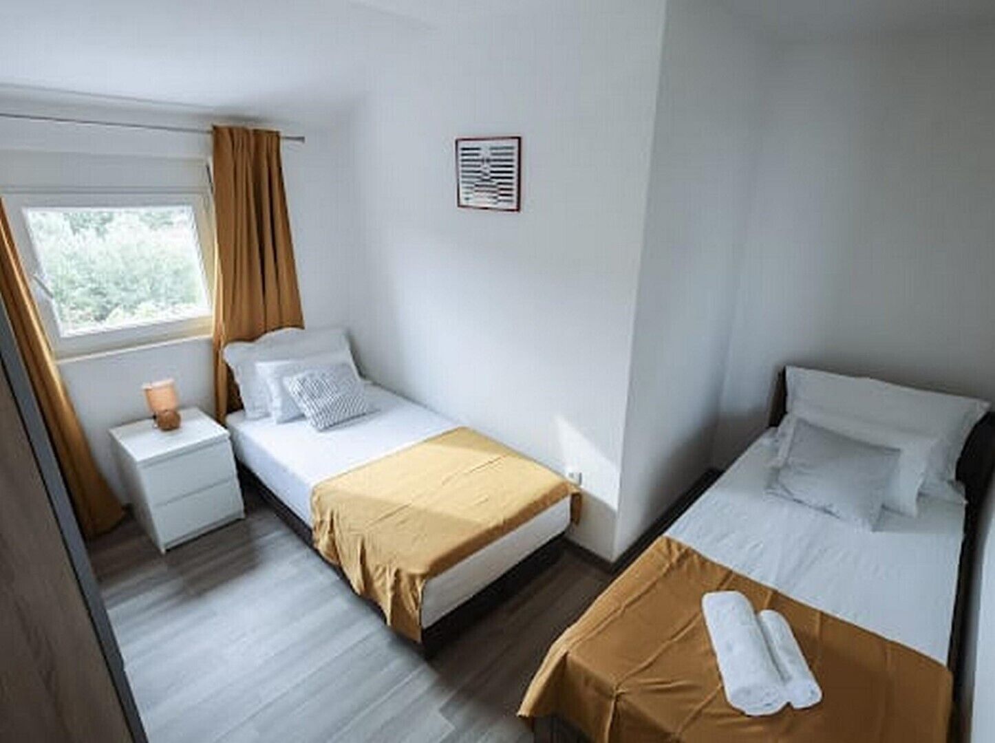 A hotel room with two single beds, a small bedside table with a lamp, a window with curtains, and a wall-mounted air conditioning unit.