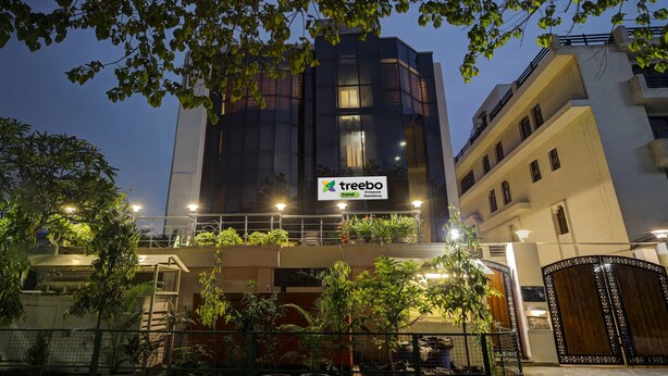 Um edifício moderno com a placa 'Treebo Trend Primeone Residency', apresentando varandas com plantas e uma entrada fechada por portão.