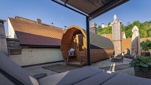 A wooden structure with a sloped roof, a small seating area with a bench and a table, and a view of the surrounding buildings and trees. - Der Postwirt (Kipfenberg)