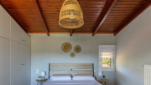 Une chambre avec un plafond en bois, un lit recouvert de draps blancs, deux tables de chevet avec des lampes et une fenêtre avec des stores.