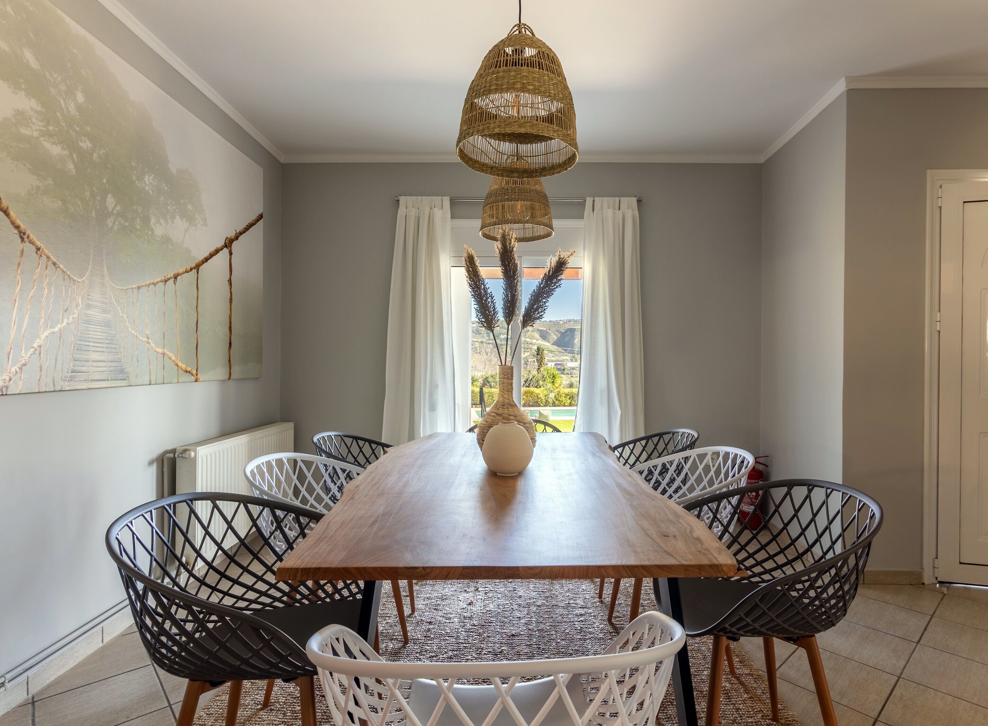 Een eethoek met een houten tafel, zes stoelen, een geweven hanglamp, een vaas met gedroogde planten en uitzicht op groen buiten het raam.