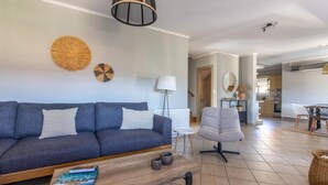 Un salon moderne avec un canapé, une table basse et un fauteuil. On aperçoit à l’arrière-plan un coin repas avec une table et des chaises.