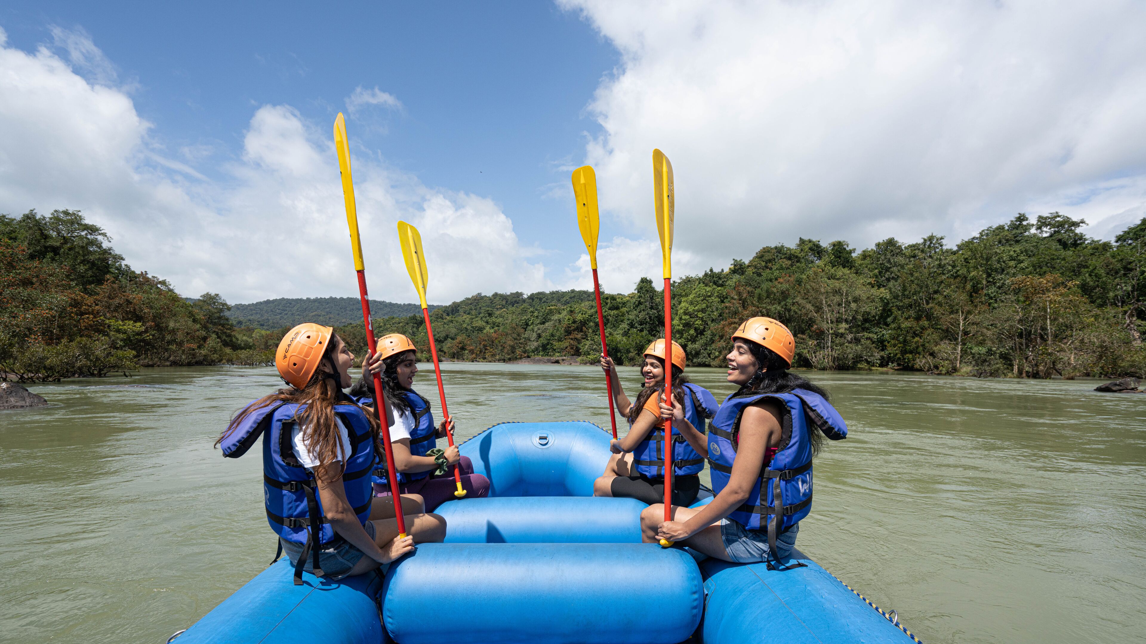 Un grupo de personas en un bote inflable azul con remos rojos, usando chalecos salvavidas y cascos, en un río rodeado de vegetación.