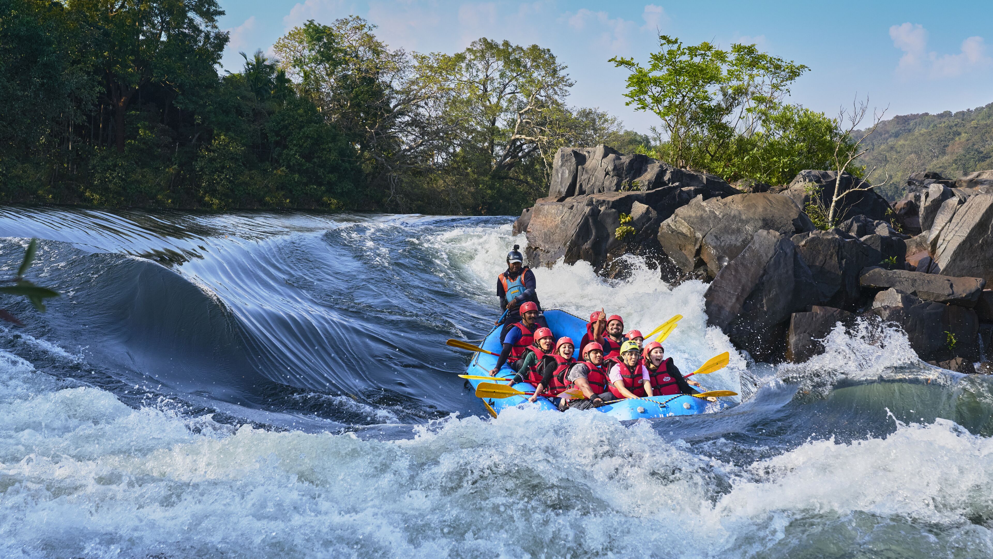 A group of people in a blue raft navigating a river with rapids, surrounded by rocky terrain and trees.