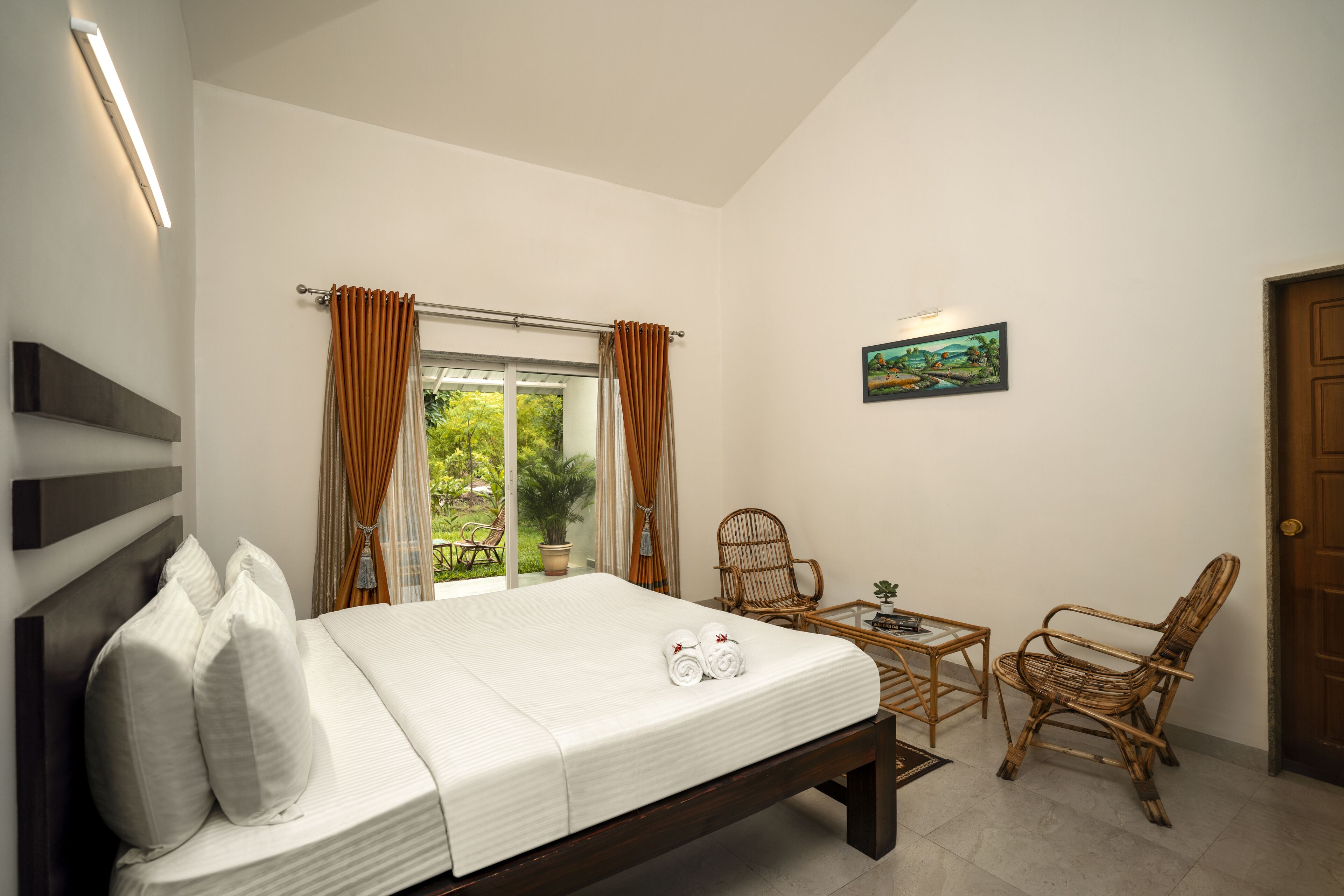 A bedroom with a bed, two wicker chairs, a small table, and a painting on the wall.