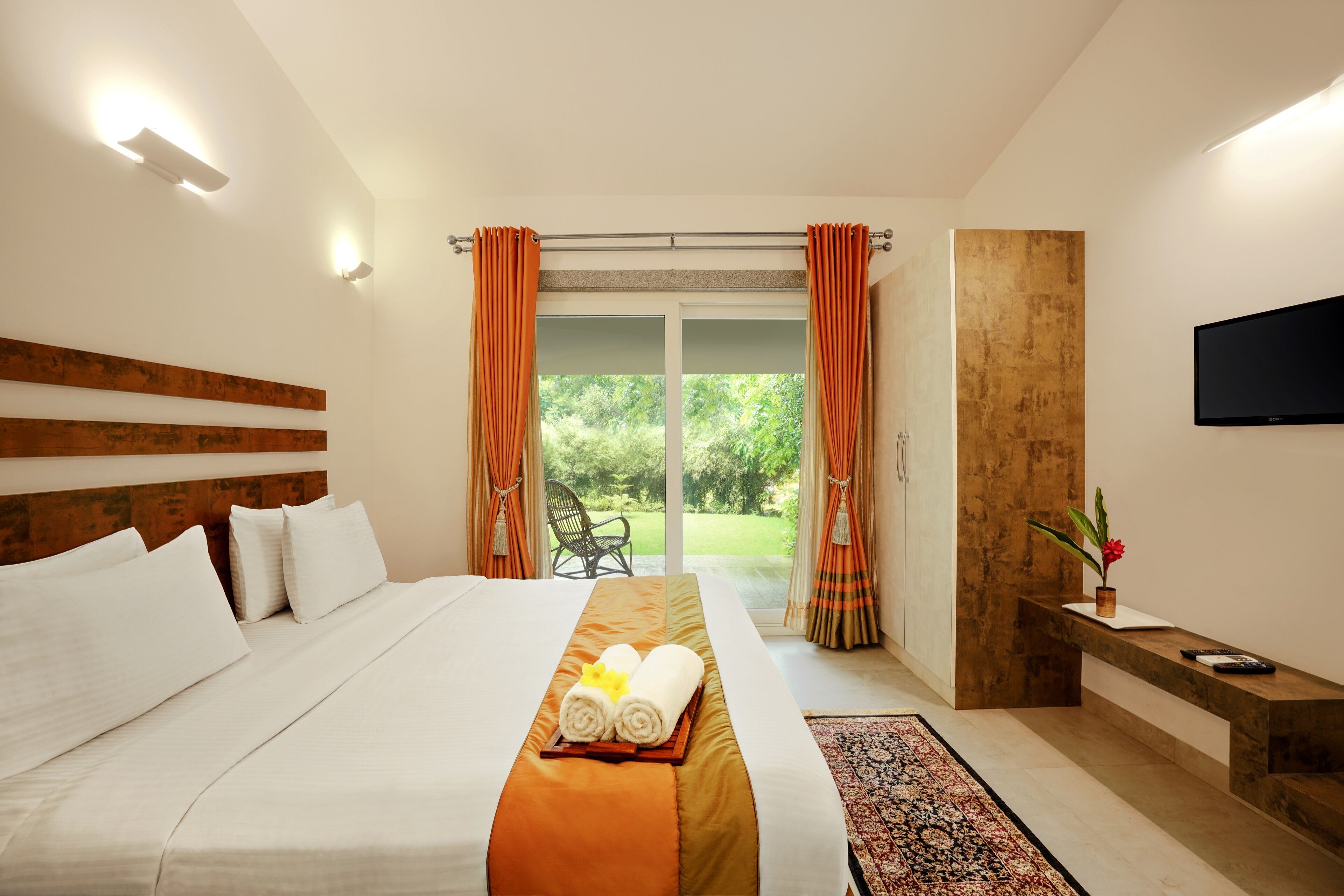 Habitación de hotel con una cama grande, cabecera de madera, televisor de pantalla plana, un banco con un florero y vistas a zonas verdes por la ventana.