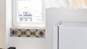 A kitchen with a white refrigerator, a window with a view of buildings, and a white pendant light. - Charming Studio in Graça Overlooking the Tagus River (Lisboa)