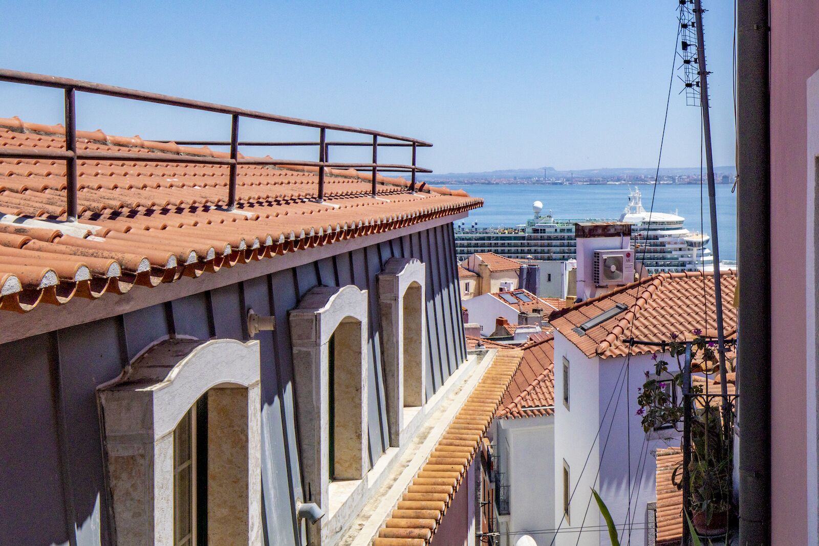 A coastal building with terracotta tiled roofs, a balcony with a metal railing, and a view of the sea and cruise ships in the distance.