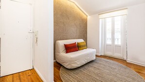 A cozy room with a white sofa, patterned wallpaper, and a large window. - Charming Studio in Graça Overlooking the Tagus River (Lisboa)