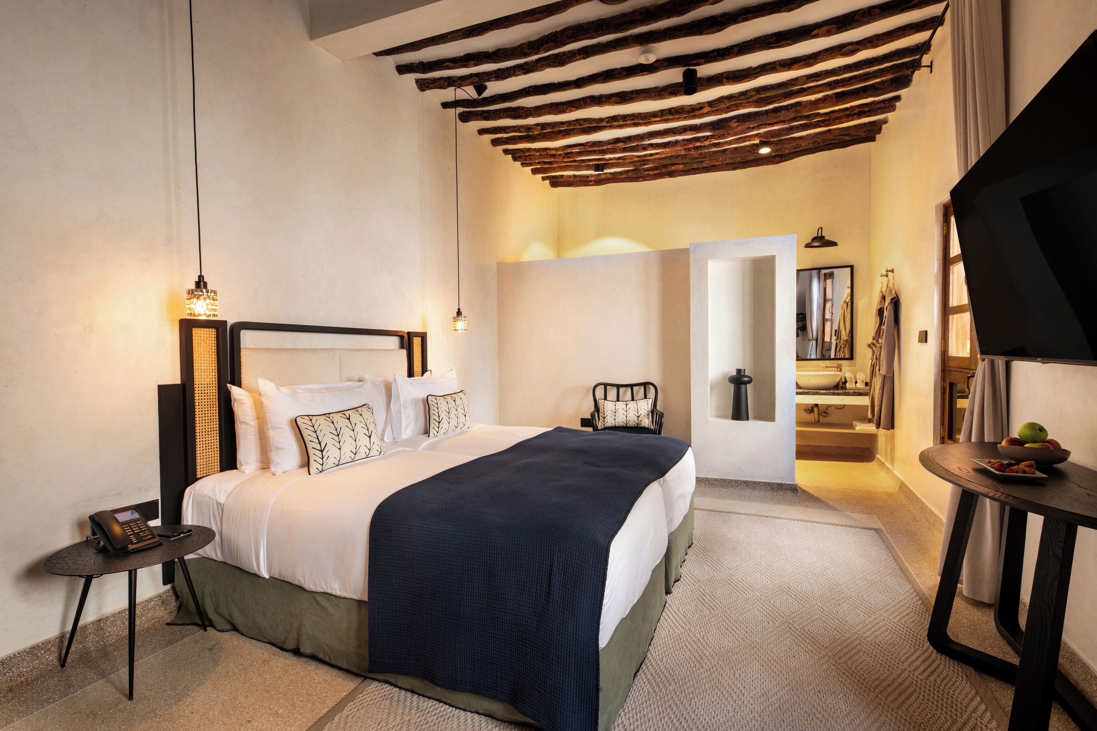 Habitación de hotel con cama, mesita de noche y una pequeña mesa con un bol de fruta.